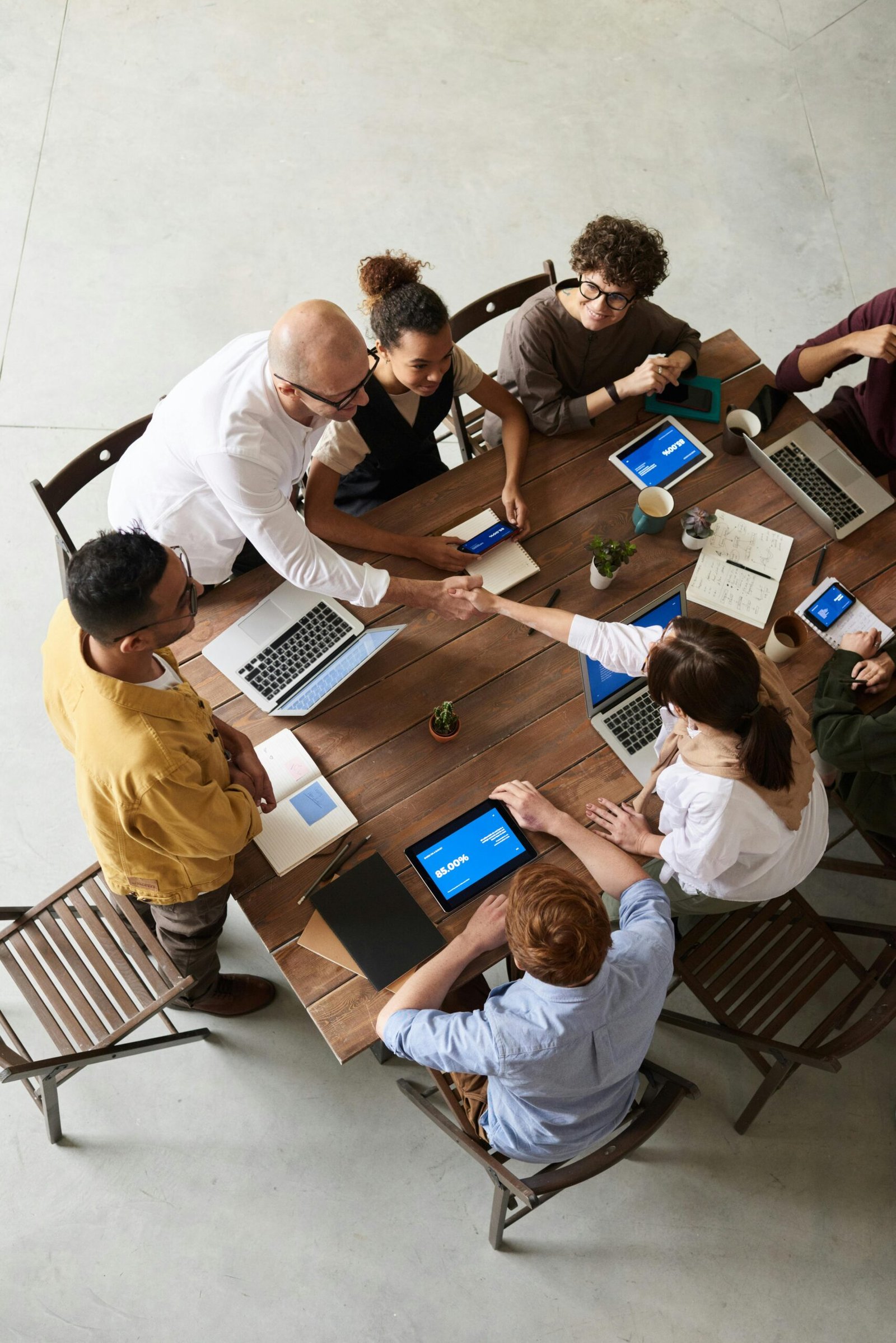 Top-down view of a group of diverse colleagues gathered around a wooden table in a collaborative meeting. Two people are shaking hands, signifying agreement or partnership, while others are engaged with laptops, tablets, and notebooks. The workspace is bright and professional, fostering teamwork and communication.